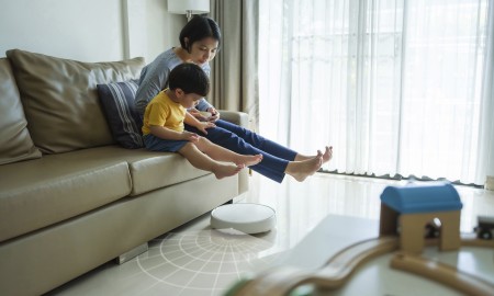 Robotic Vacuum Cleaner Cleaning Floor while Asian Japanese Mother relaxing with her little son on sofa in the living room. Woman and boy lift feet up together