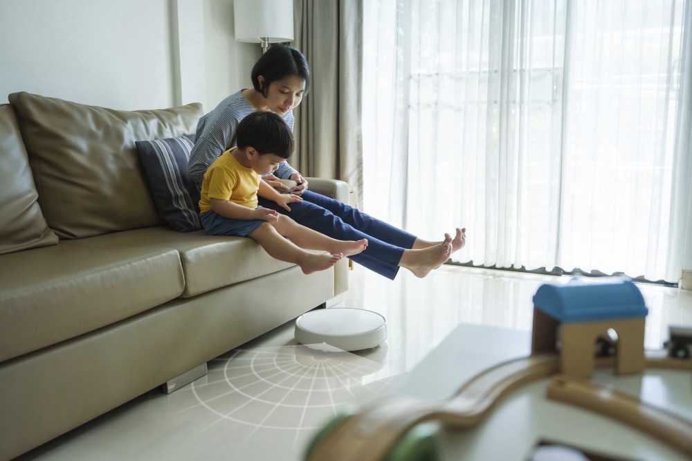 Robotic Vacuum Cleaner Cleaning Floor while Asian Japanese Mother relaxing with her little son on sofa in the living room. Woman and boy lift feet up together